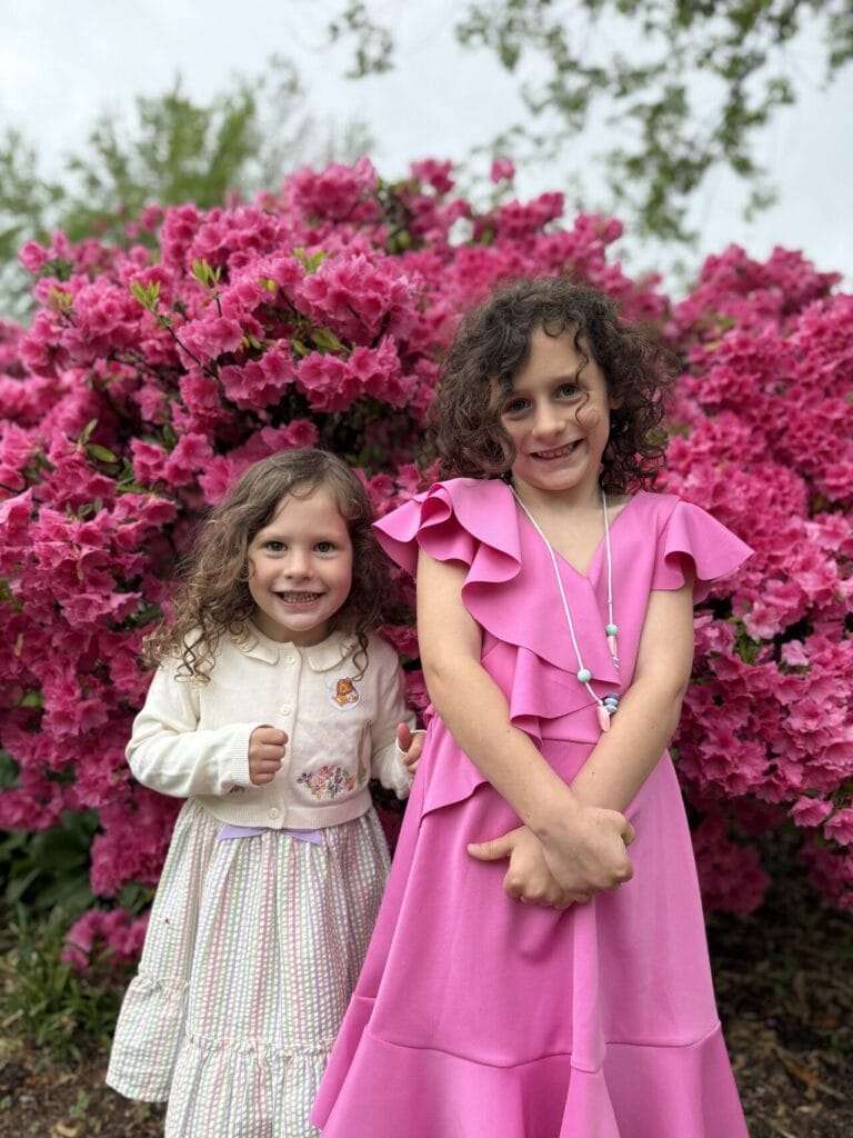 Two young girls in Easter dresses smiling in front of a large pink azalea bush