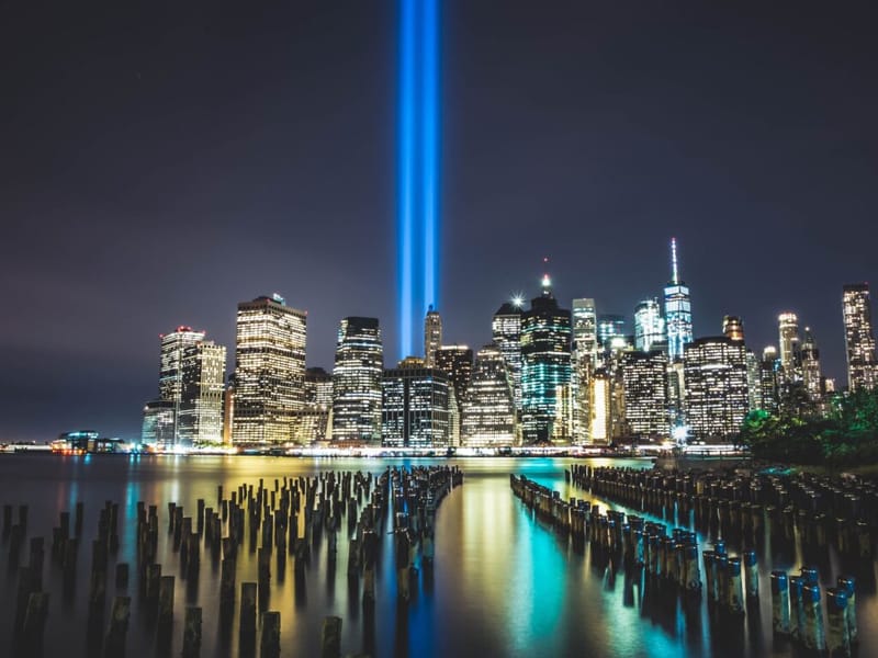 Tribute in Light memorial beams rising above the Lower Manhattan skyline at night, reflected in the water, with old wooden pier pilings in the foreground