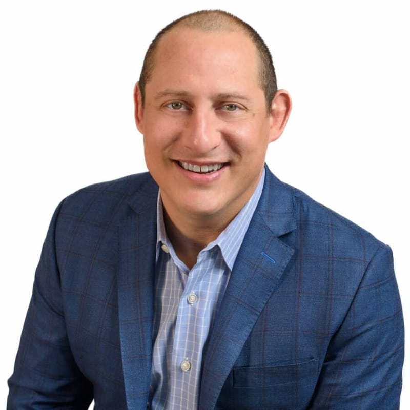 Adam Buice headshot in a blue plaid blazer, smiling against a white background