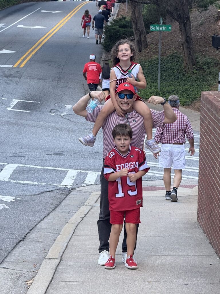 Adam Buice with his daughter on his shoulders and son in a Georgia Bulldogs jersey, walking toward a game on Baldwin Street