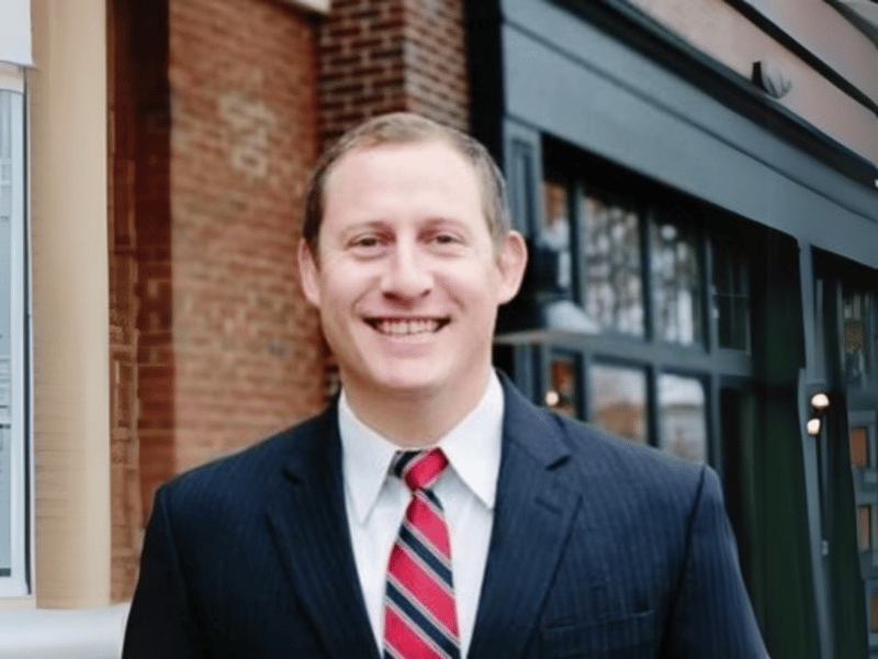 Adam Buice smiling outdoors in a navy blazer and striped tie, standing in front of a brick building