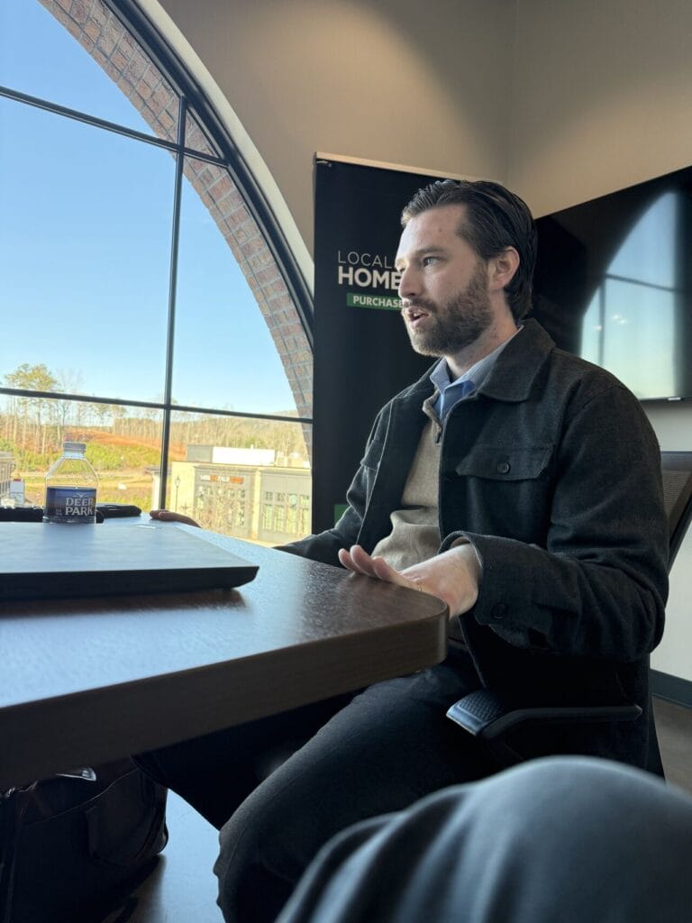 Adam Buice seated at a table during a meeting, speaking with hands clasped, Local Home Purchasers banner and large arched window visible behind him