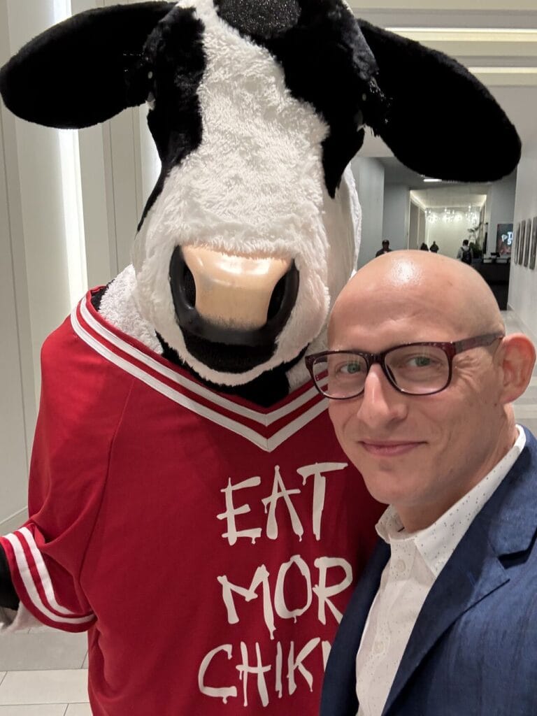 Adam Buice posing with the Chick-fil-A cow mascot in a red "Eat Mor Chikin" jersey at an indoor venue
